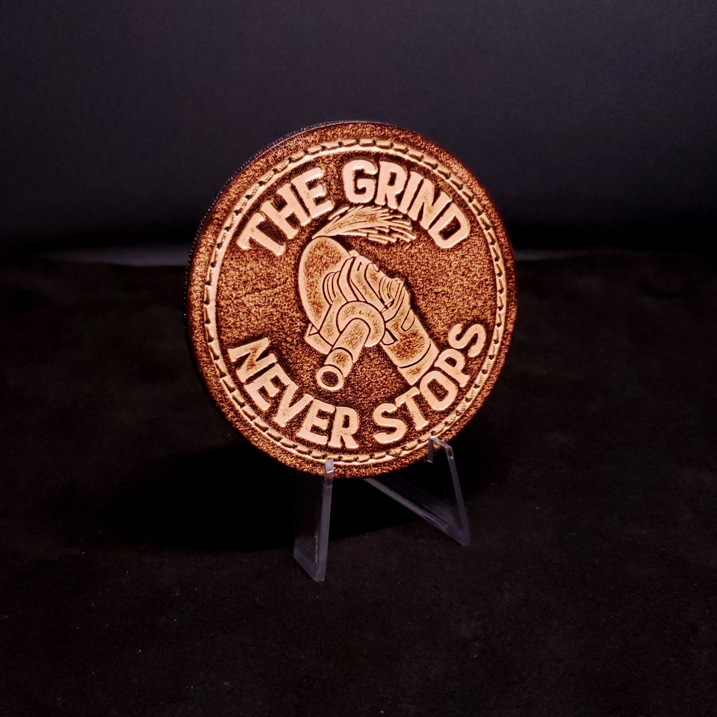 Brown leather velcro patch with text that says "The grind never stops" and an image of an angle grinder, posed on a black background.