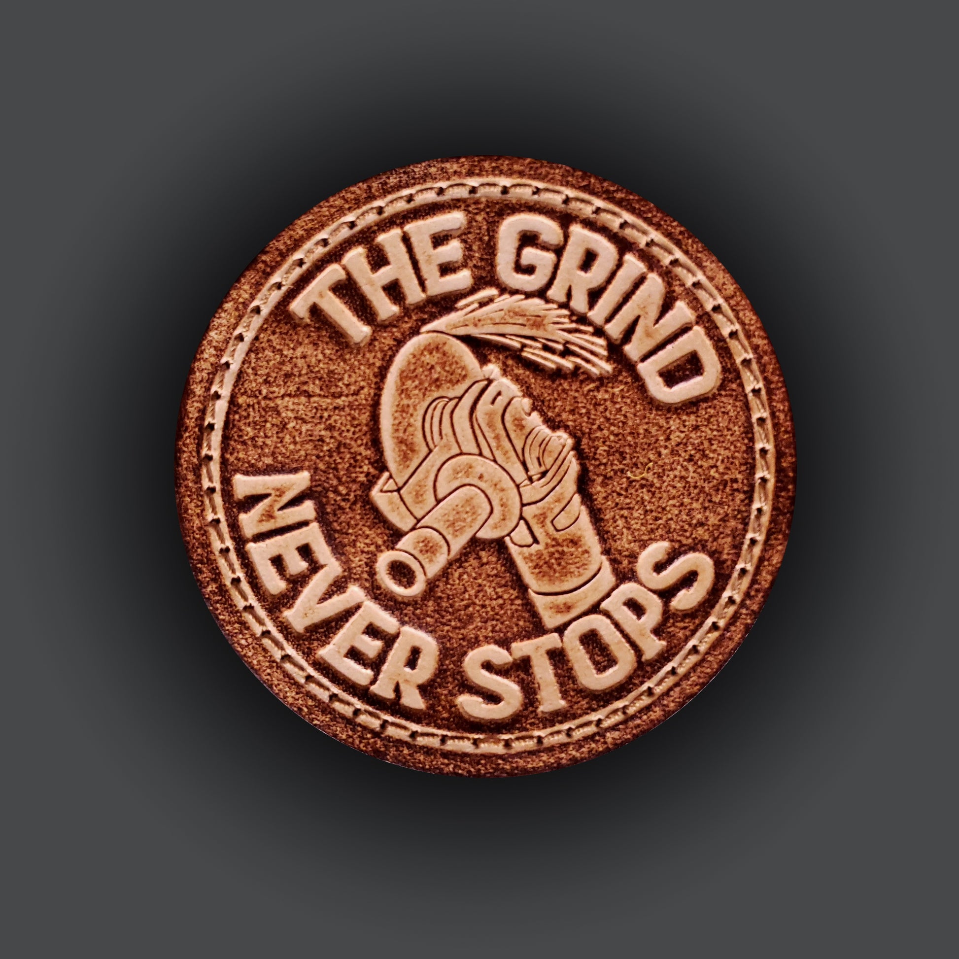 Brown leather velcro patch with text that says "The grind never stops" and an image of an angle grinder and sparks.