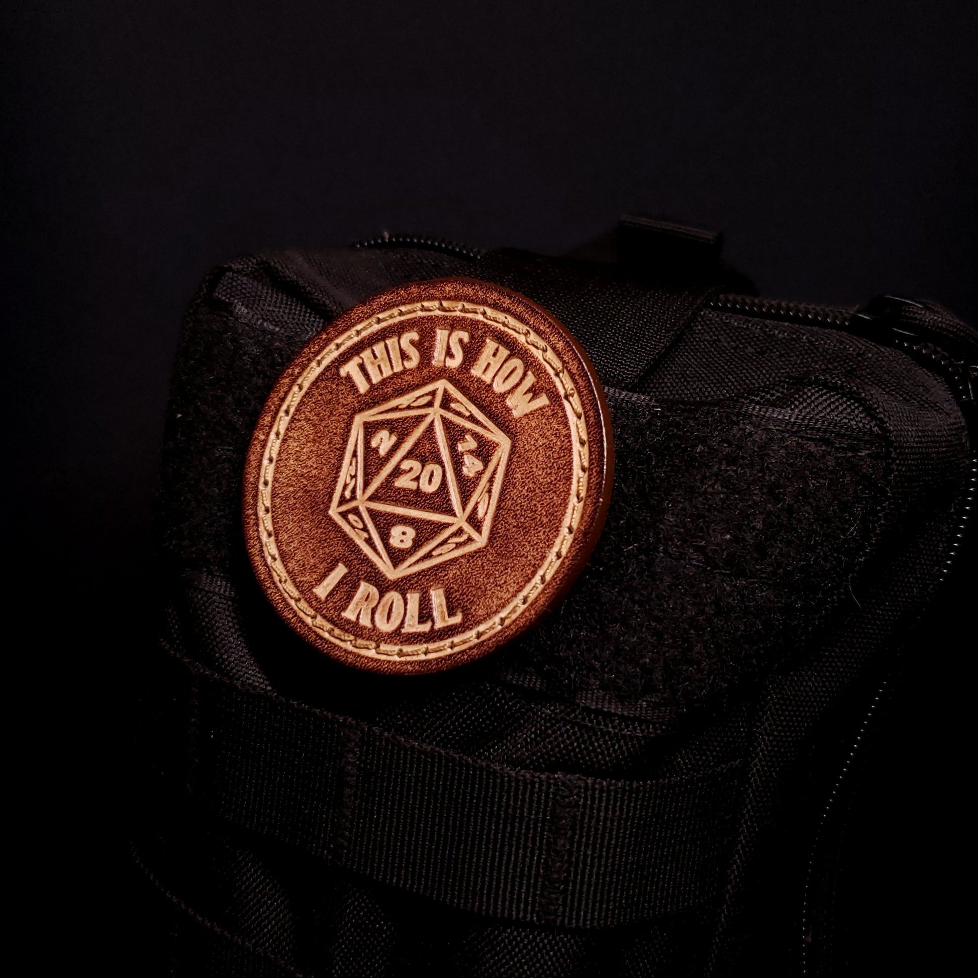 Brown leather velcro patch with text and an image of a twenty sided die with the number twenty facing up, affixed to a bag.