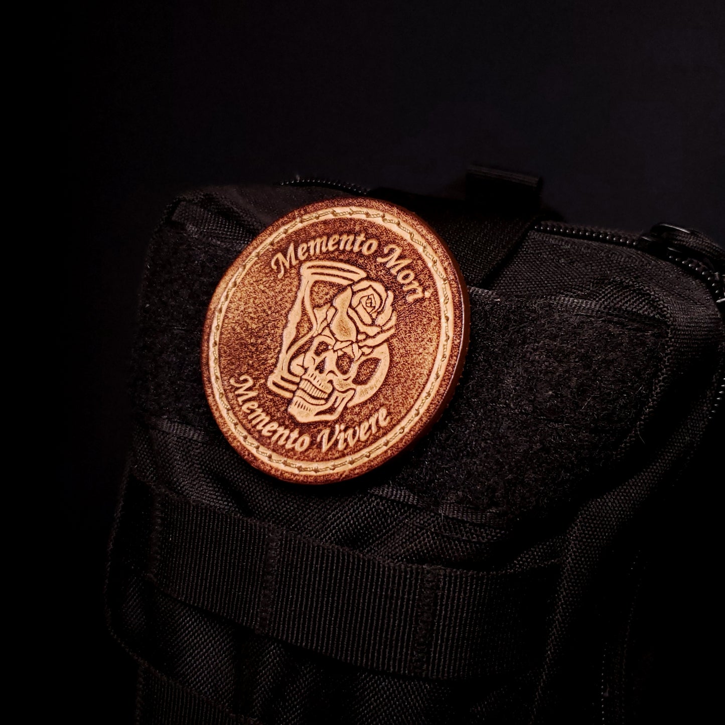 Brown leather velcro patch with text that says an image of a skull, rose, and hourglass, affixed to a bag.