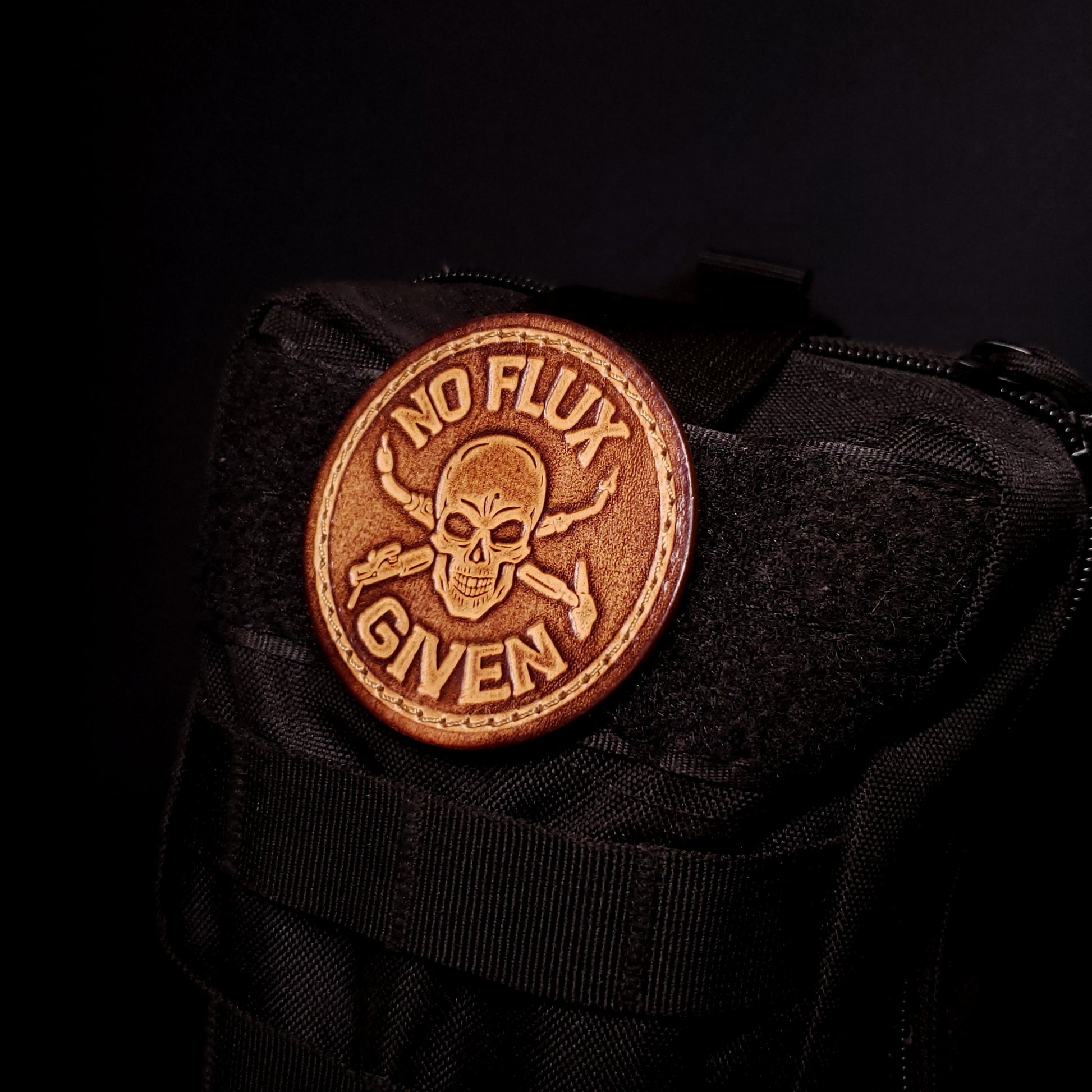 Brown leather velcro patch embossed with text that says "No flux given", and an image of a skull with welding tools crossed behind it, affixed to a bag.