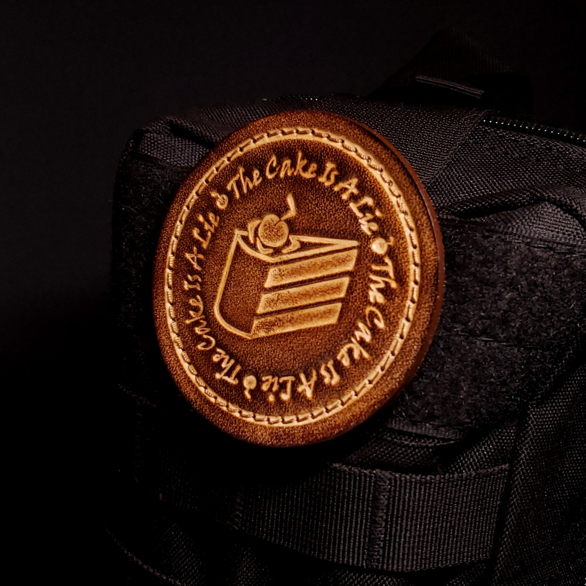 Brown leather velcro patch embossed with text and a graphic of cake from the Portal franchise, affixed to a bag.