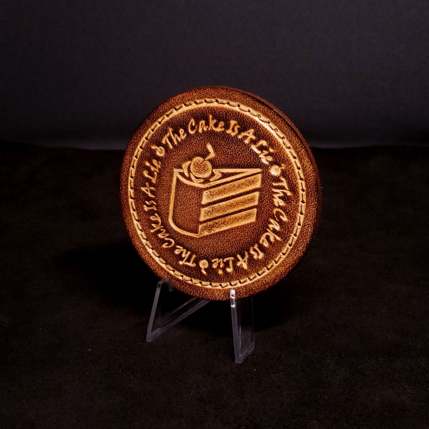 Brown leather velcro patch embossed with text and a graphic of cake from the Portal franchise, posed on a black background.