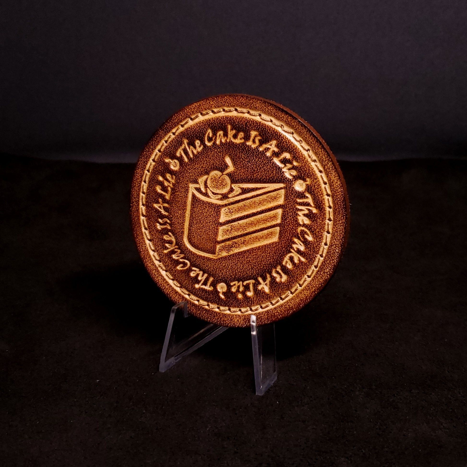 Brown leather velcro patch embossed with text and a graphic of cake from the Portal franchise, posed on a black background.