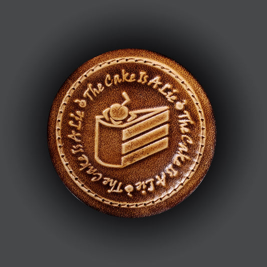 Brown leather velcro patch embossed with text that says "The cake is a lie", and a piece of cake from the Portal franchise.