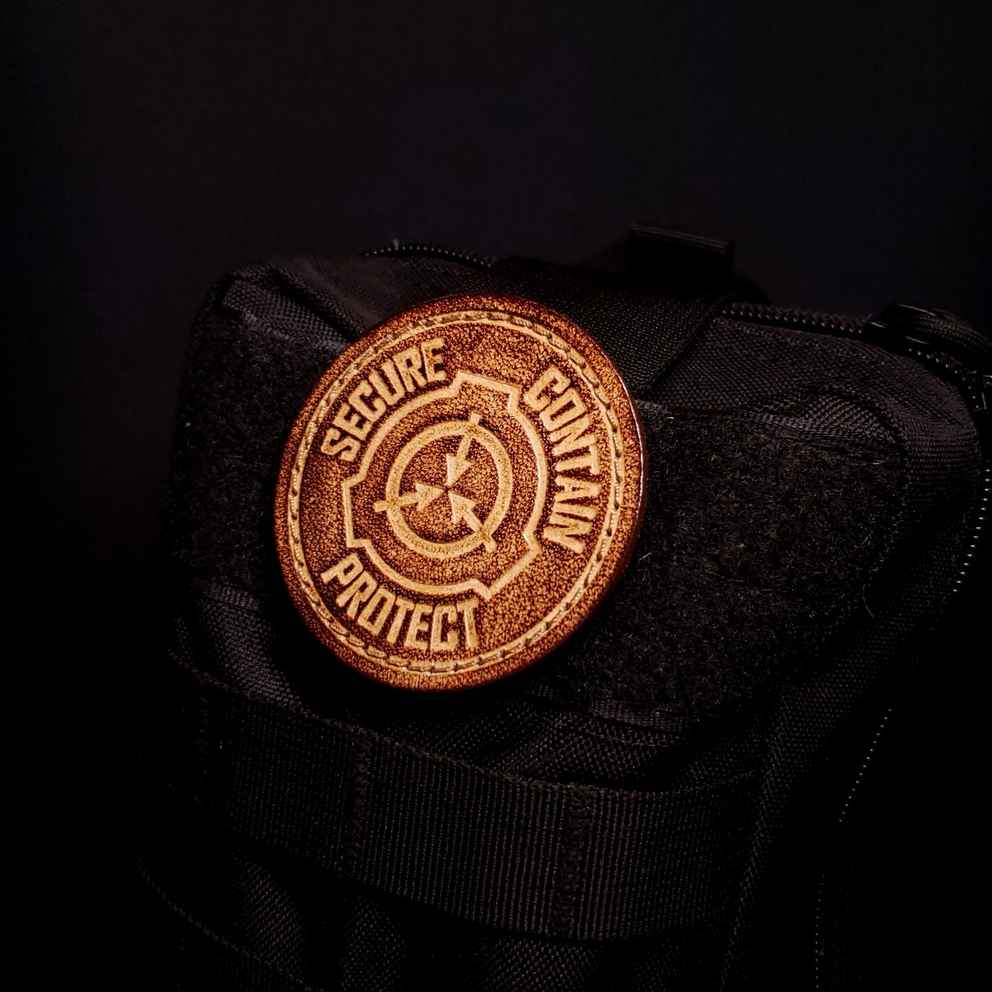 Brown leather velcro patch embossed with text that says "Secure, contain, protect", and a symbol of the fictional SCP foundation, affixed to a bag.