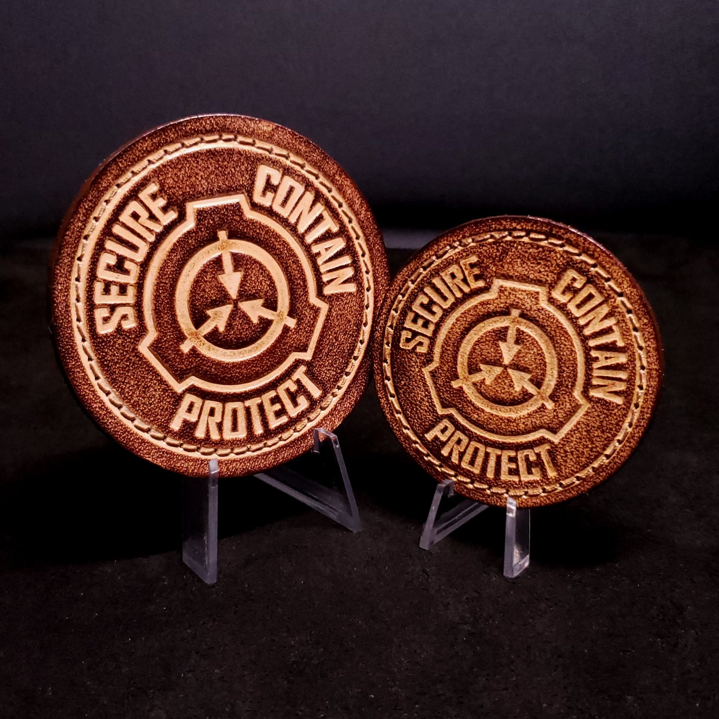 Two brown leather velcro patches embossed with text that says "Secure, contain, protect", and a symbol of the fictional SCP foundation, posed on a black background.